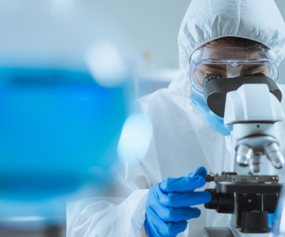 Osoba w białym kombinezonie i goglach patrzy przez mikroskop. O walce z wirusem ebola przeczytasz na Radioplus.