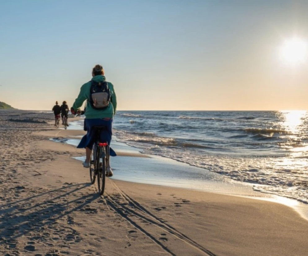 Osoby jadące na rowerach po piaszczystej plaży nad morzem o zachodzie słońca. O udanym urlopie przeczytasz w Radioplus.