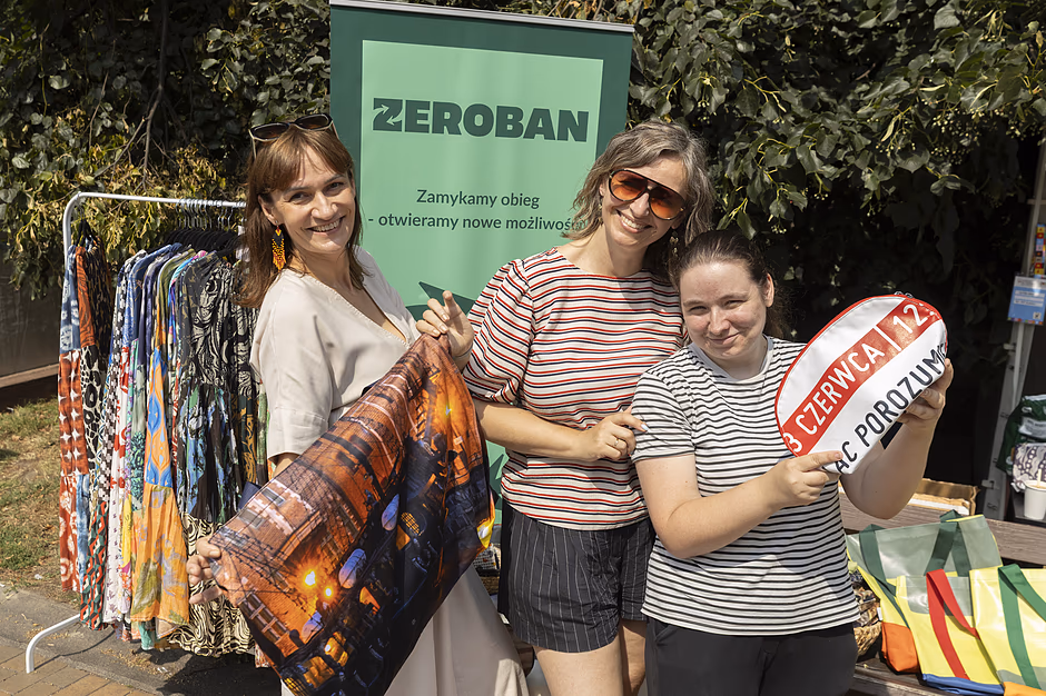 trzy kobiety pozują, pokazując torby, za nimi baner z napisem Zeroban oraz wieszak z długimi kolorowymi spódnicami