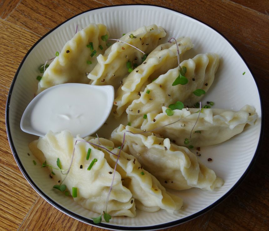 Na talerzu znajdują się pierogi, posypane ziołami, obok miseczka z jogurtem.