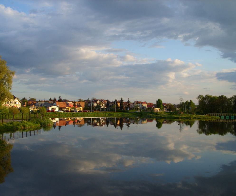 Widok na zalew w Kazimierzy Wielkiej, w którym odbijają się domy i chmury. O nowym uzdrowisku przeczytasz w Eska Kielce.