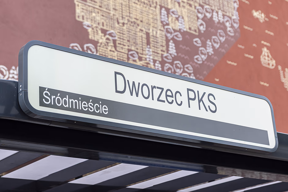 Zdjęcie przedstawia białą tablicę informacyjną umieszczoną na konstrukcji wiaty przystankowej. Widnieją na niej czarne napisy wskazujące nazwę przystanku „Dworzec PKS” oraz dzielnicę „Śródmieście” na ciemniejszym pasku.