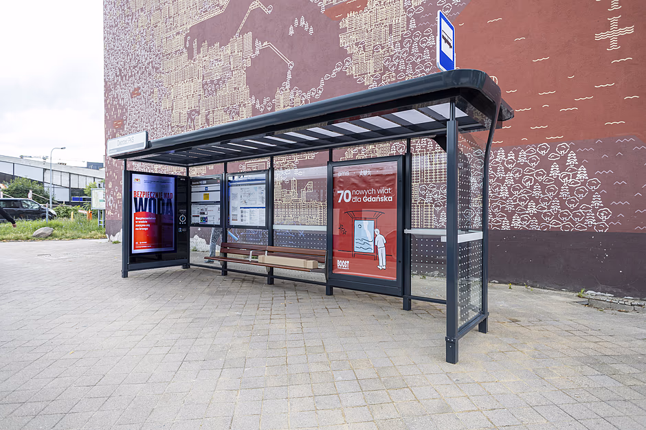 Zdjęcie przedstawia nowoczesną wiatę autobusową ustawioną na tle dużej ściany budynku pokrytej brązowym muralem. Konstrukcja przystanku jest wyposażona w podświetlane gabloty reklamowe, rozkład jazdy oraz drewnianą ławkę dla pasażerów.