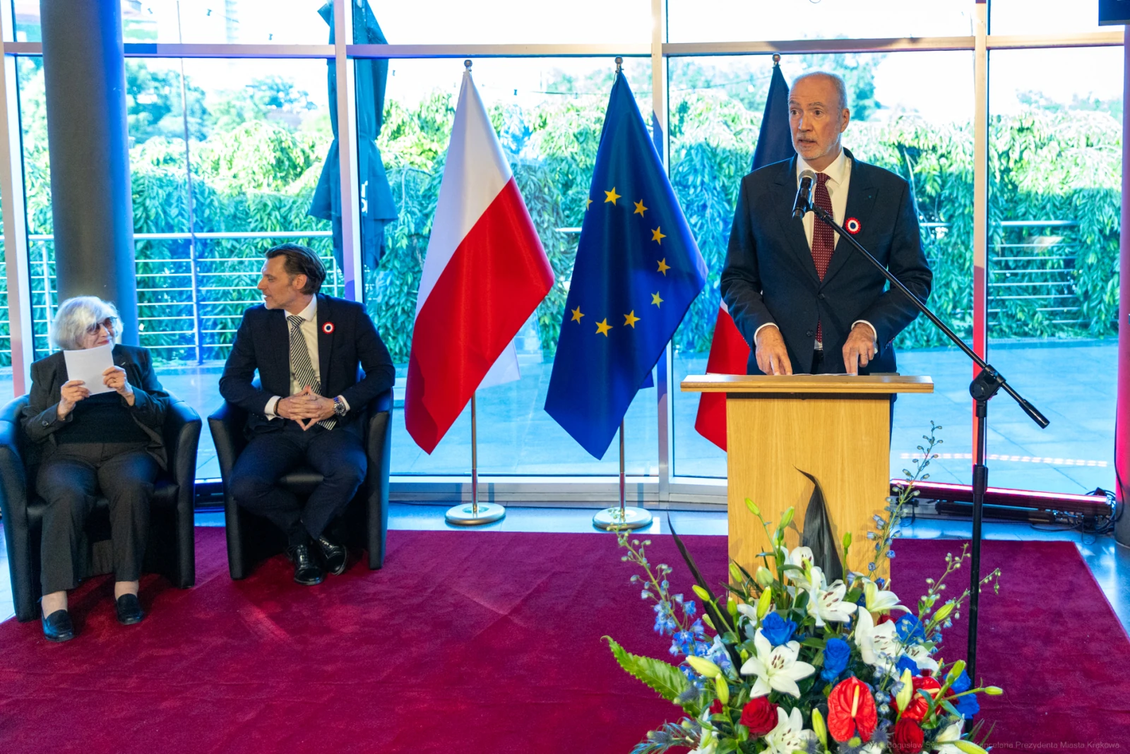 Mężczyzna przemawia przy mównicy, obok siedzą dwie osoby, w tle flagi Polski i Unii Europejskiej. Autor: Bogusław Świerzowski
