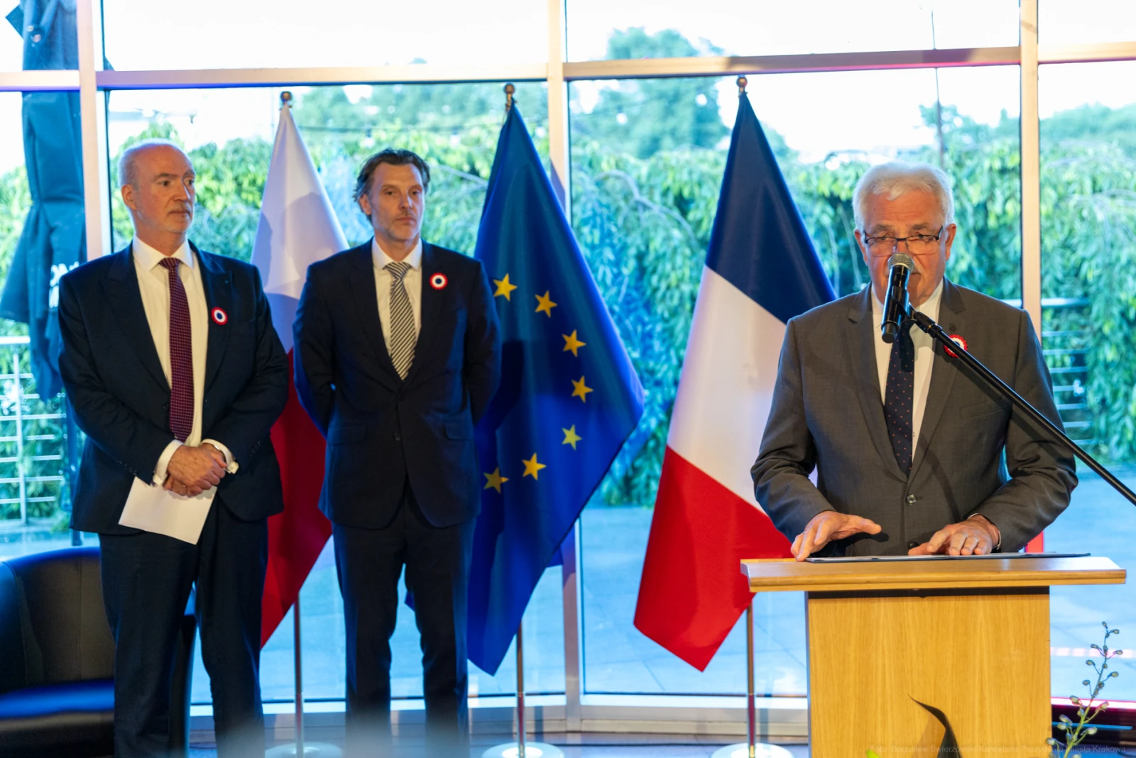 Mężczyzna przemawia przy mównicy, obok dwóch mężczyzn w garniturach, w tle flagi Polski, UE i Francji. Autor: Bogusław Świerzowski