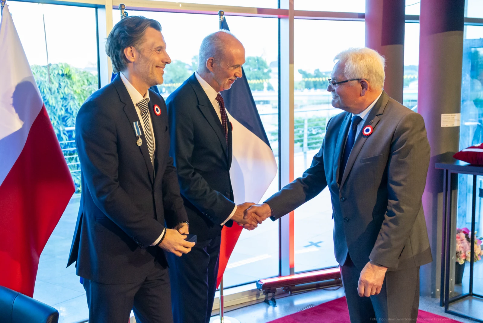 Trzech mężczyzn w garniturach, uściski dłoni, flagi Polski i Francji w tle. Autor: Bogusław Świerzowski