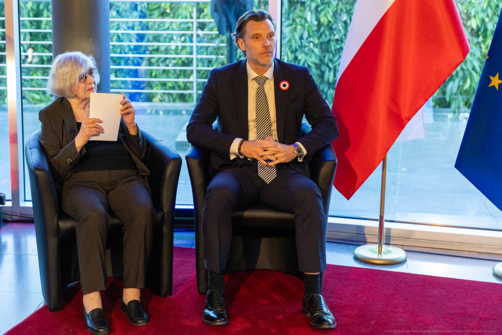 Dwoje ludzi siedzących na kanapie, obok flagi Polski i Unii Europejskiej. Autor: Bogusław Świerzowski