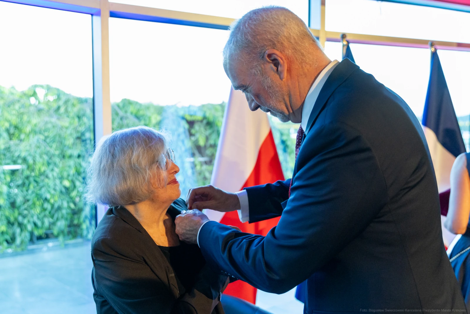 Wręczenie odznaczenia kobiecie przez mężczyznę w eleganckim otoczeniu. Autor: Bogusław Świerzowski