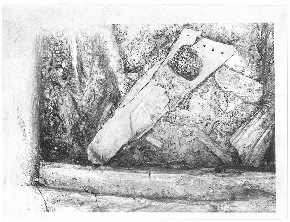 Czarno-biała fotografia przedstawia zbliżenie na podłoże archeologiczne z widocznymi fragmentami drewna oraz kamieni. W centralnej części kadru wyróżnia się podłużny, płaski element drewniany ze śladami otworów, częściowo przysypany ziemią.