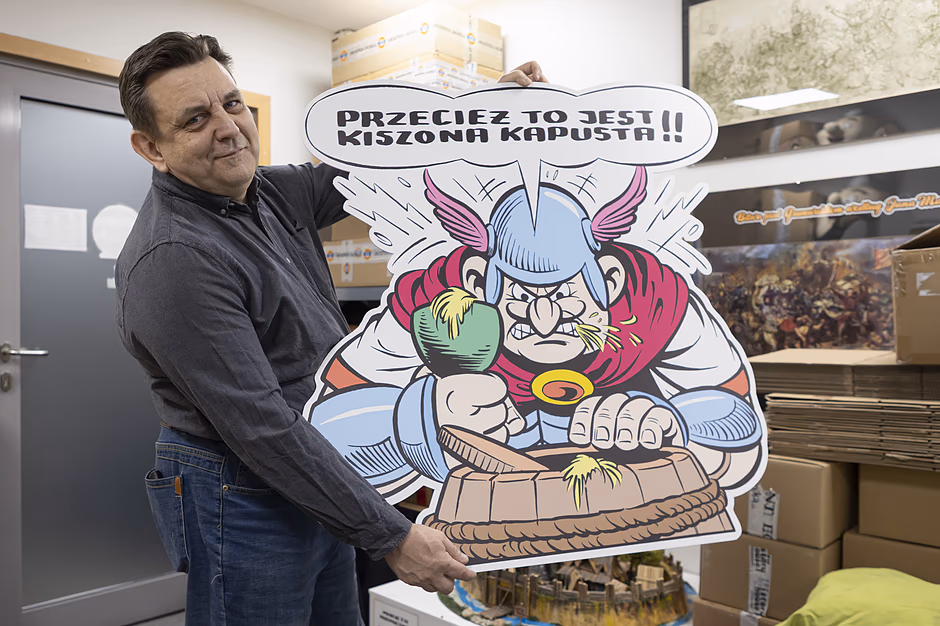 Mężczyzna w ciemnej koszuli trzyma dużą, komiksową wycinankę przedstawiającą wściekłego Gala w rogatym hełmie. Postać na planszy uderza pięścią w drewnianą beczkę, a nad nią widnieje komiksowy dymek z napisem „PRZECIEŻ TO JEST KISZONA KAPUSTA!!”.