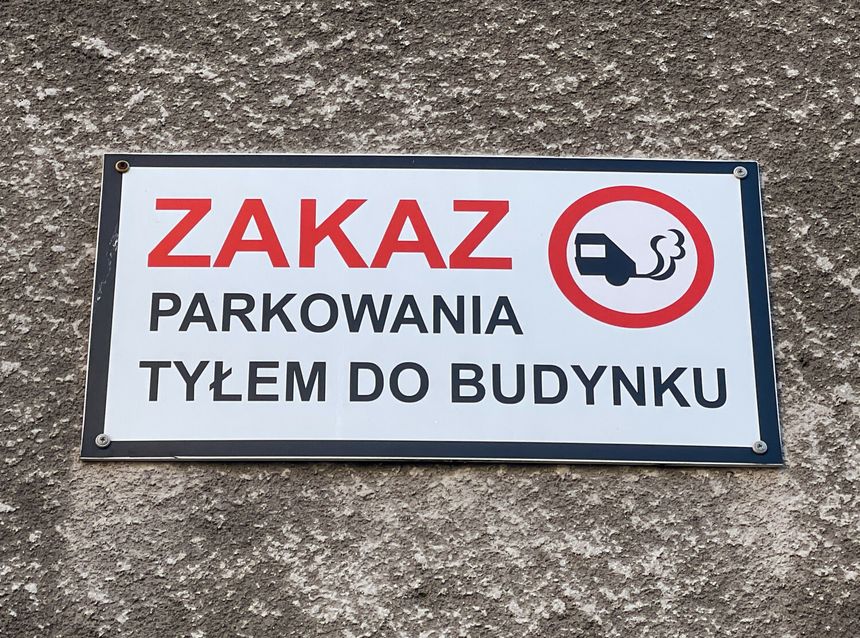 Znak zakazu parkowania tyłem do budynku, z symbolem ciężarówki z dymem.