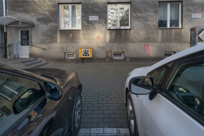 Widok na szare ściany budynku, między dwoma zaparkowanymi samochodami, z graffiti i znakami.