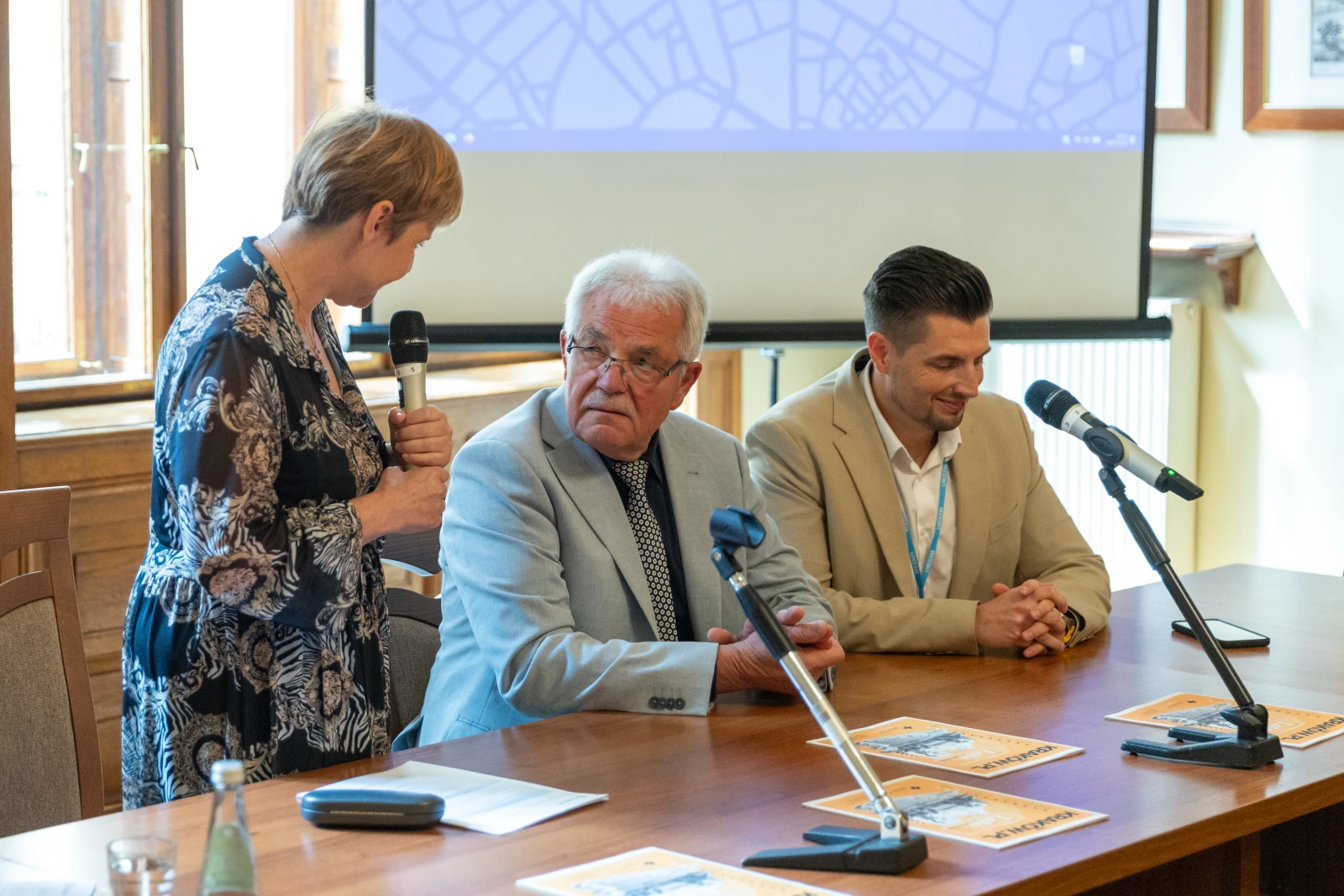 Spotkanie trzech osób przy stole, jedna z nich mówi do mikrofonu, w tle ekran. Autor: Bogusław Świerzowski