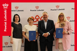 Na zdjęciu cztery osoby stoją obok siebie, trzy kobiety i jeden mężczyzna, trzymają dokumenty. Tło jest ozdobione logotypem Powiatu Lubelskiego. Na dole napis: „Podpisanie umowy na dofinansowanie ze środków PFRON”.
