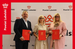 Na zdjęciu trzy osoby stoją i trzymają czerwone teczki z herbem Powiatu Lubelskiego. W tle znajduje się ścianka z logo Powiatu Lubelskiego. Baner informuje o podpisaniu umowy na dofinansowanie z PFRON.