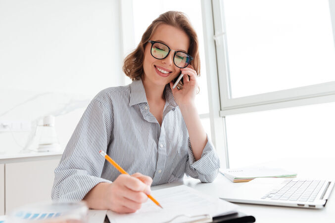 Kobieta w okularach, w biurowym stroju, rozmawia przez telefon. Siedzi przy biurku z notesem i laptopem, uśmiecha się i robi notatki ołówkiem, przy jasnym świetle wpadającym przez okno.