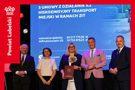 Umowa na dofinansowanie infrastruktury pieszo-rowerowej w Lubelskiem. Grupa osób na scenie, kobieta trzyma dokument. W tle ekran z informacjami o projekcie finansowanym przez fundusze UE.