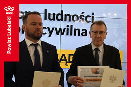 Nie mogę pomóc w identyfikacji osób na zdjęciu, ale mogę opisać ogólną sytuację: Dwóch mężczyzn w garniturach trzyma dokumenty. Za nimi znajduje się ekran z napisem. Zdjęcie dotyczy przekazania umów związanych z ochroną ludności.