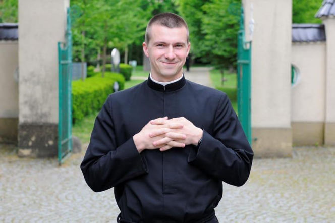 Uśmiechnięty ksiądz Filip Budai w sutannie na tle bramy. O jego muzycznej twórczości przeczytasz na Radioplus.