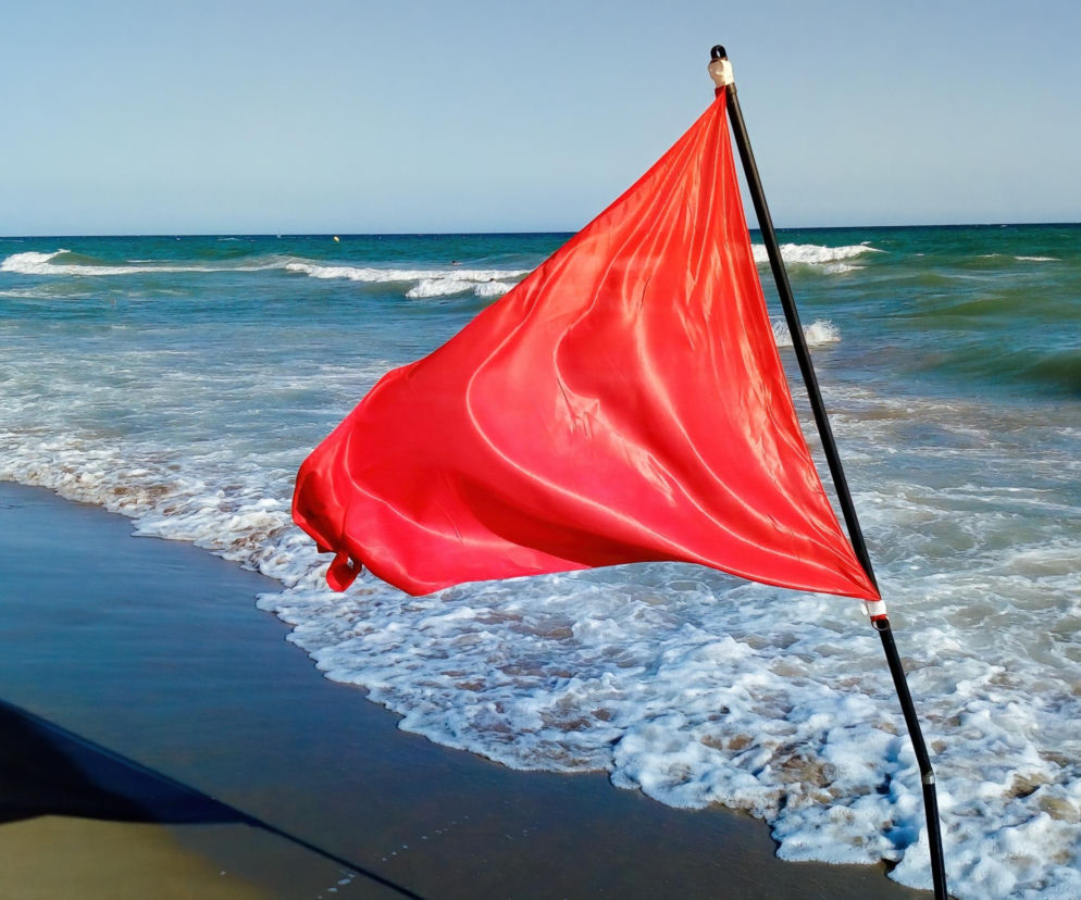 Czerwona flaga powiewająca na plaży przy wzburzonym morzu. O mandatach dla turystów przeczytasz na Eska Trójmiasto.