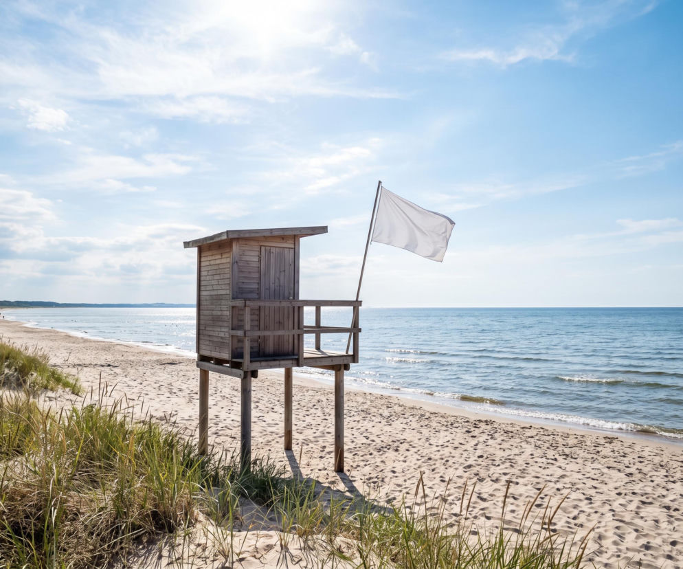 Drewniana wieżyczka ratownicza z białą flagą na pustej, piaszczystej plaży. O pogodzie w Dąbkach przeczytasz na SE Szczecin.