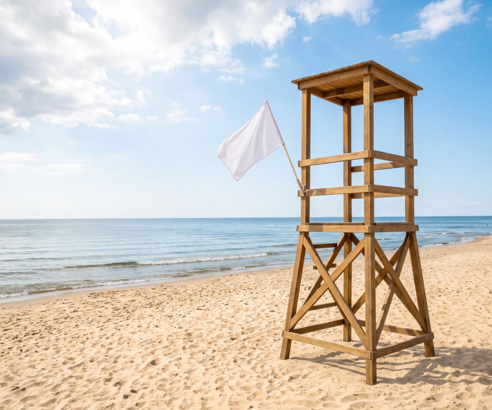 Drewniana wieża ratownicza z białą flagą na pustej, piaszczystej plaży. O pogodzie w Dziwnowie przeczytasz w SE Szczecin.