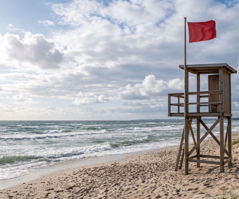 Wieża ratownicza z czerwoną flagą na plaży przy wzburzonym morzu. O warunkach w Kołobrzegu przeczytasz na SE Szczecin.