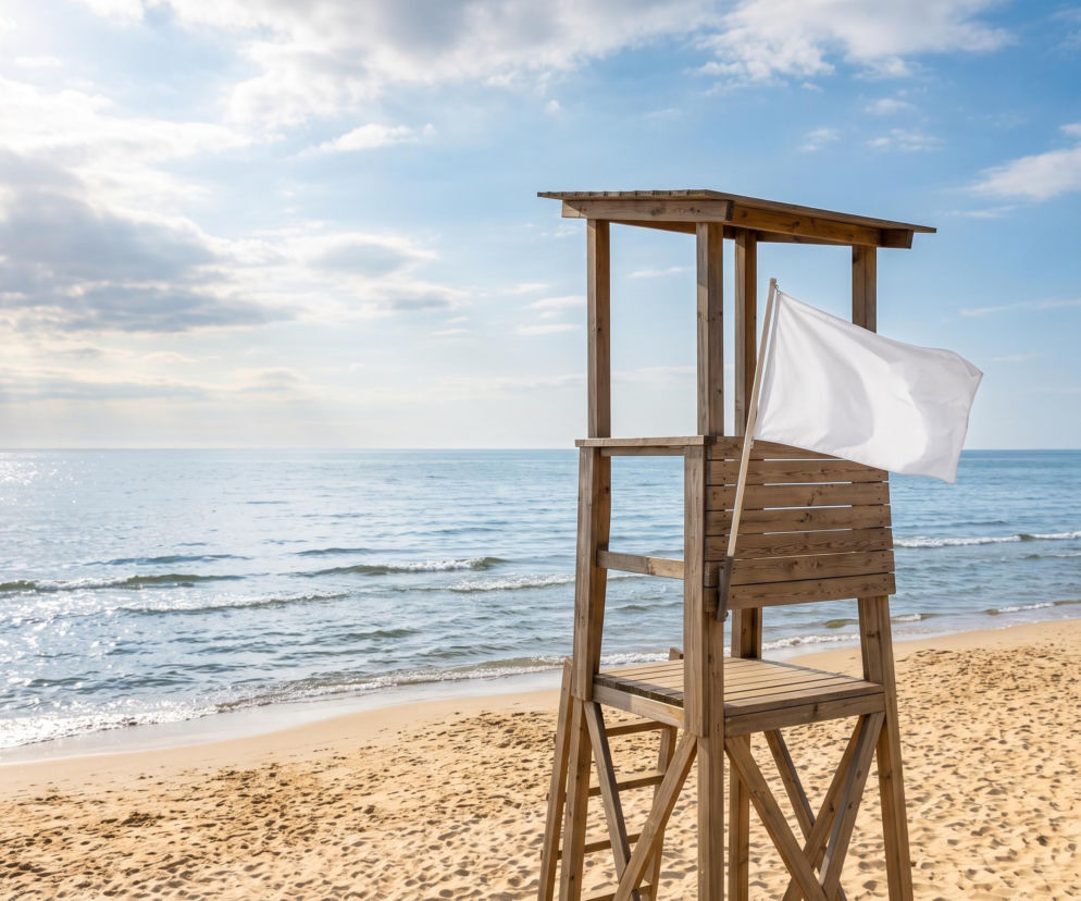 Drewniana wieża ratownika na piaszczystej plaży z powiewającą białą flagą. O warunkach w Krynicy przeczytasz na Eska Trójmiasto.