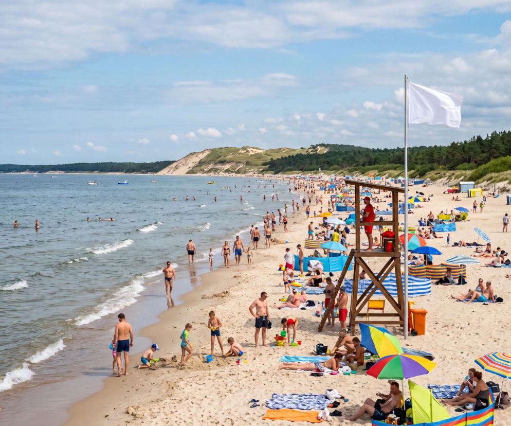 Ludzie na słonecznej plaży w Łebie. Obok wieży ratownika powiewa biała flaga. O bezpieczeństwie przeczytasz w Eska Trójmiasto.