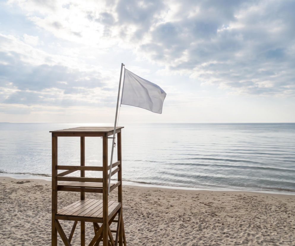 Biała flaga na wieży ratowniczej przy pustej plaży i spokojnym morzu. O braku sinic w Rewalu przeczytasz na SE Szczecin.