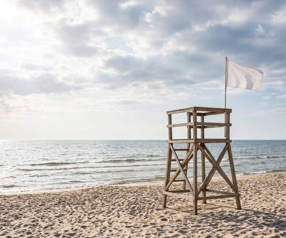 Drewniana wieża ratownicza na piaszczystej plaży z białą flagą. O warunkach kąpieli przeczytasz na Eska Trójmiasto.