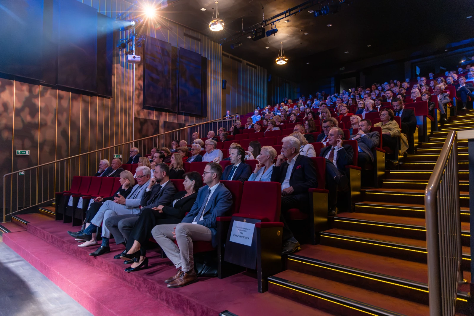 Widownia w teatrze z siedzącymi widzami, oświetlenie sceny i czerwone fotele. Autor: Bogusław Świerzowski