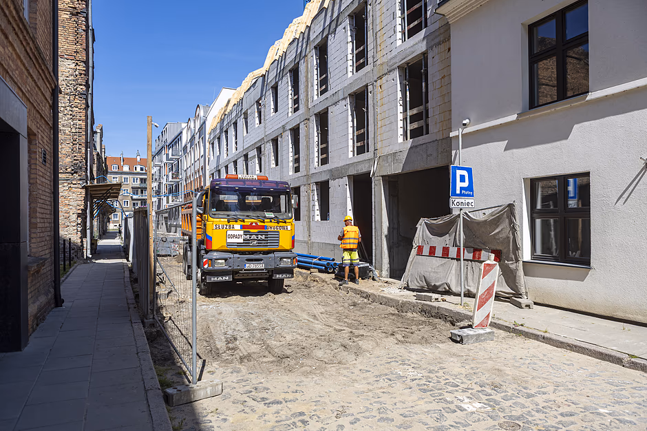 Fotografia przedstawia plac budowy nowego budynku wielorodzinnego zlokalizowanego wzdłuż wąskiej, brukowanej ulicy miejskiej. Na ogrodzonym terenie stoi pomarańczowa ciężarówka marki MAN, obok której pracuje robotnik w odblaskowej kamizelce i kasku ochronnym.