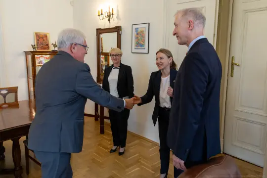 Spotkanie dwóch osób przy uścisku dłoni, w tle kobieta i mężczyzna w formalnych strojach.