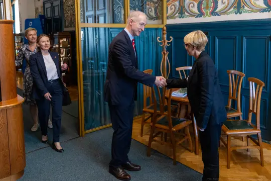 Spotkanie dwóch osób przy uścisku dłoni w eleganckim wnętrzu. W tle inne osoby.