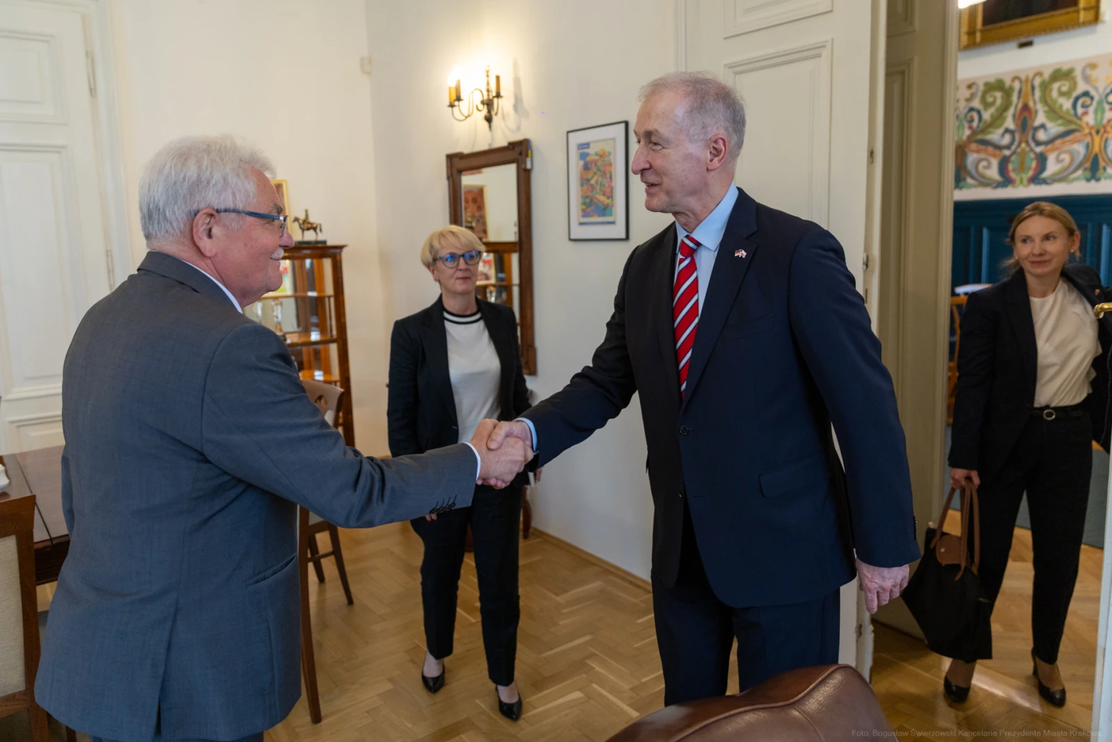Mężczyzna w garniturze podaje rękę innemu mężczyźnie, w tle kobiety w formalnych strojach. Autor: Bogusław Świerzowski