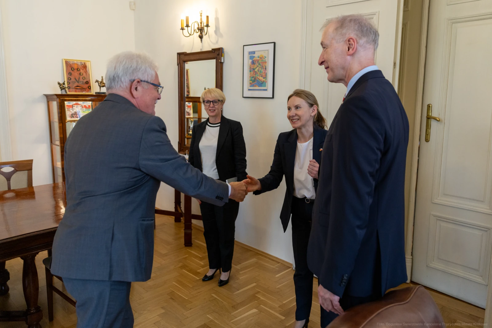 Spotkanie dwóch osób przy uścisku dłoni, w tle kobieta i mężczyzna w formalnych strojach. Autor: Bogusław Świerzowski