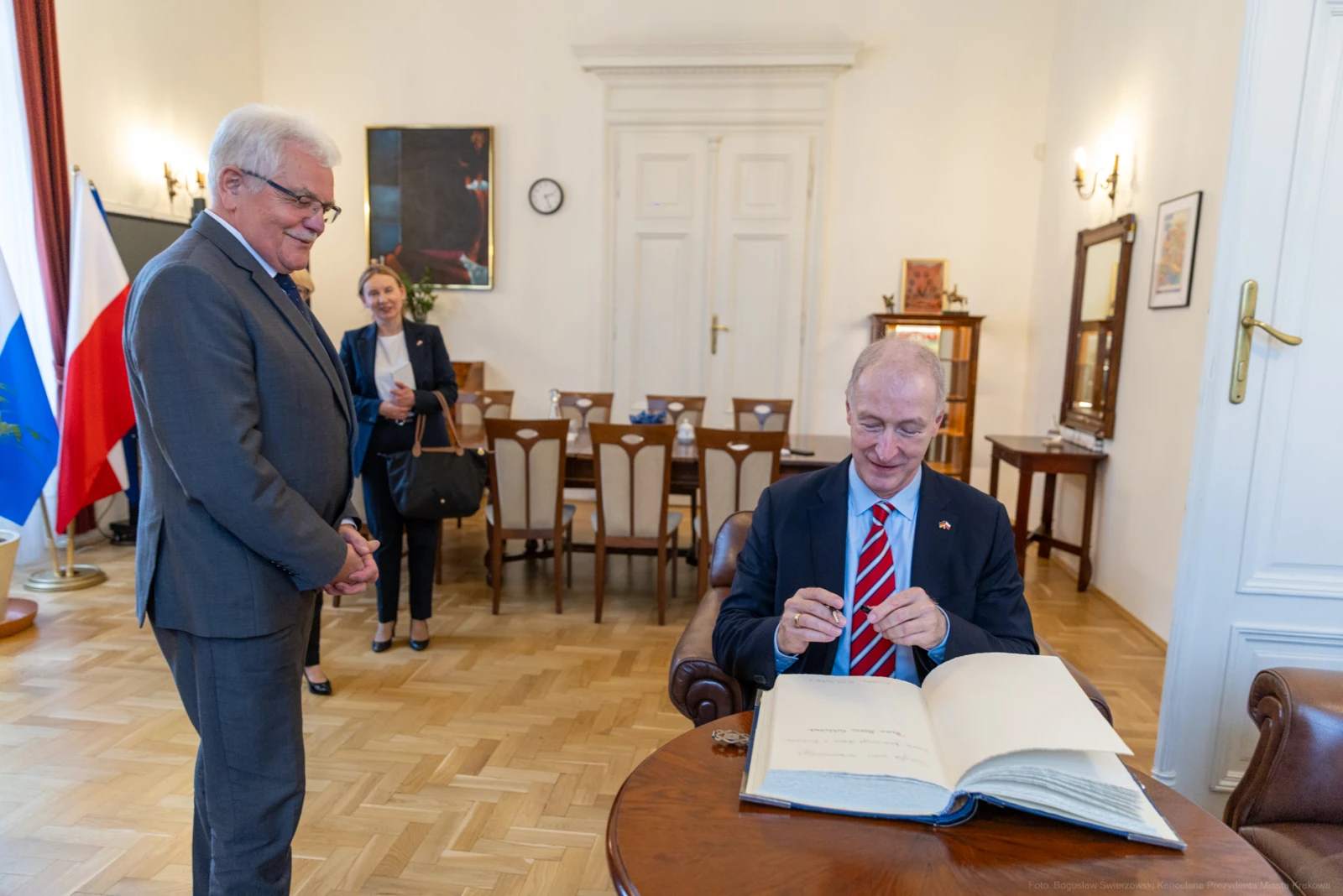 Mężczyzna w garniturze podpisuje księgę, obok stoi inny mężczyzna i kobieta. Autor: Bogusław Świerzowski